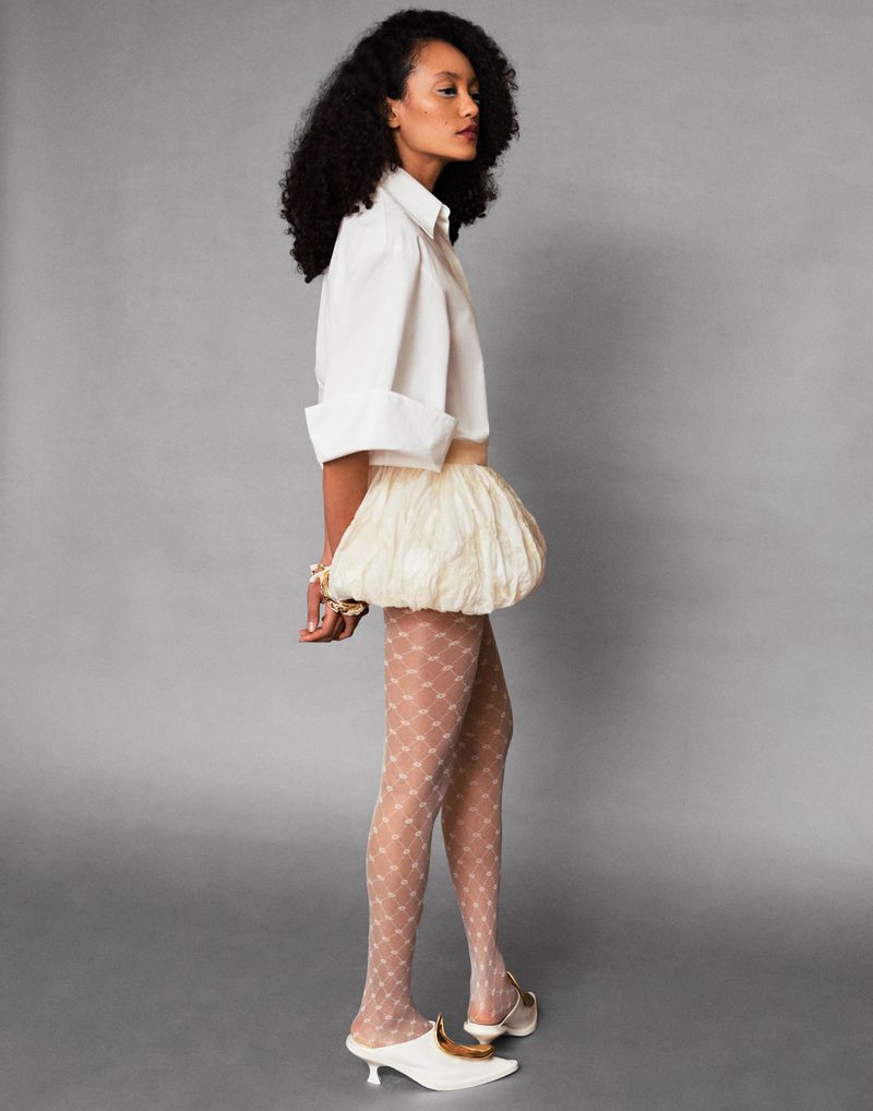 Model in a white collared shirt, cream bubble skirt, patterned tights, and kitten heels standing against a grey studio background.