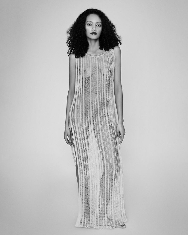 Full-length black and white fashion portrait of a woman wearing a sheer floor-length knit dress against a plain studio background.