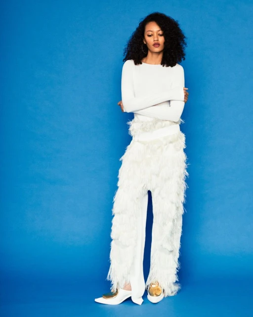 Woman in white top and feathered pants poses against blue backdrop in a fashion shot, looking down with arms crossed in studio lighting.