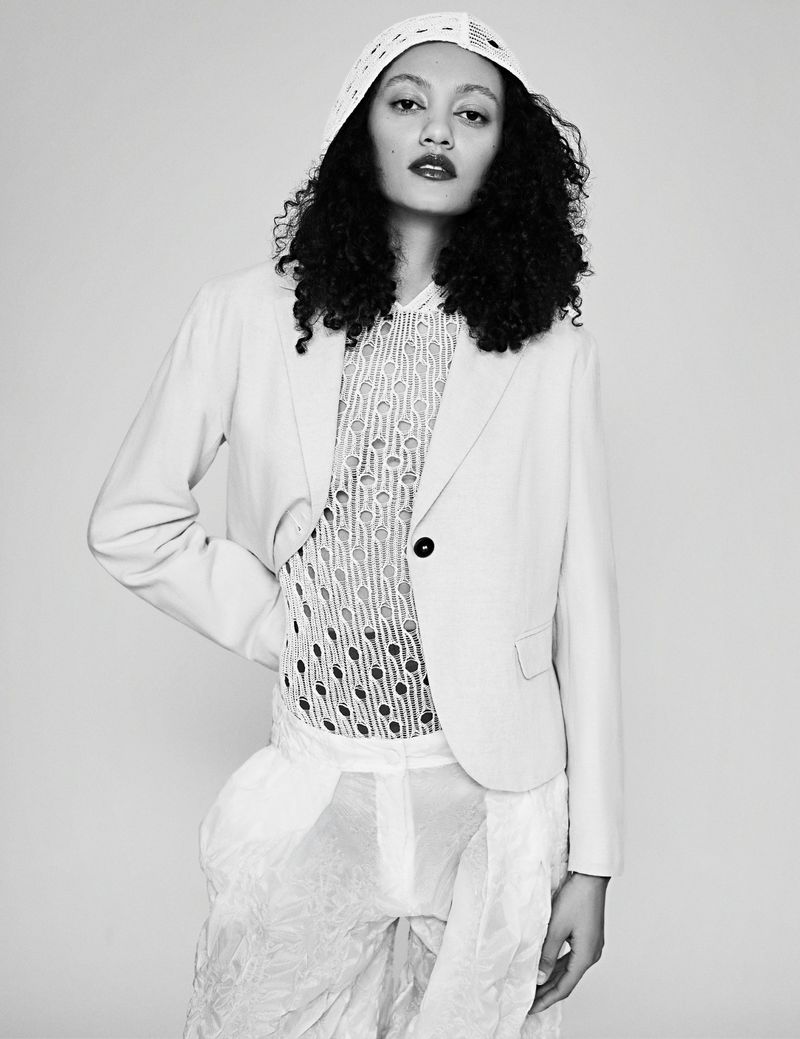 Monochrome portrait of a model with dark curly hair wearing a blazer, sheer shirt, and textured pants, set against a light background.