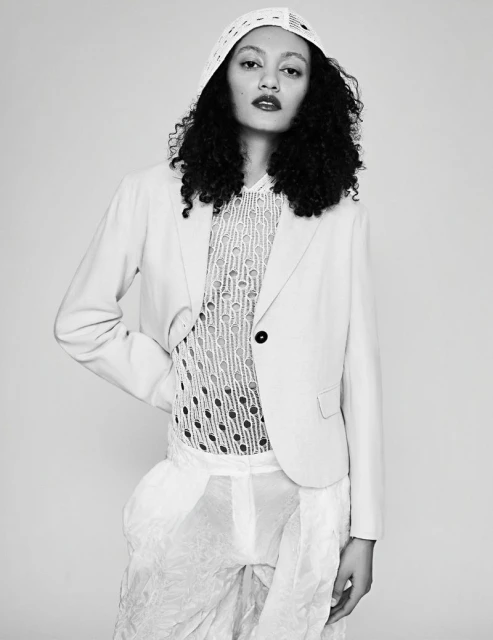 Monochrome portrait of a model with dark curly hair wearing a blazer, sheer shirt, and textured pants, set against a light background.