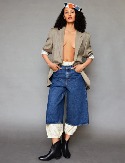 Woman in oversized blazer, blue culottes, and ankle boots poses in a studio with a colorful hat. Fashion editorial.