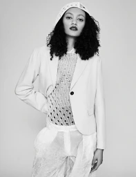 Monochrome studio portrait of a woman wearing a tailored jacket over a textured mesh top and hooded headpiece.