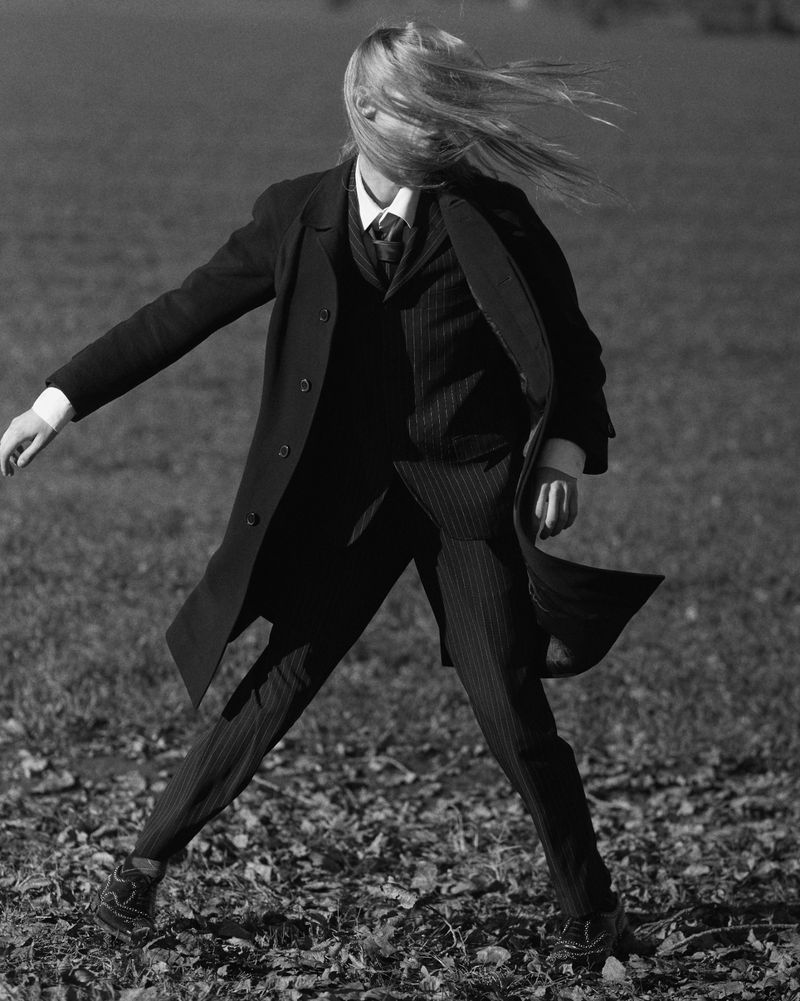 Woman in suit and coat with windblown hair stands on a field covered in fallen leaves in a black and white outdoor fashion shot.