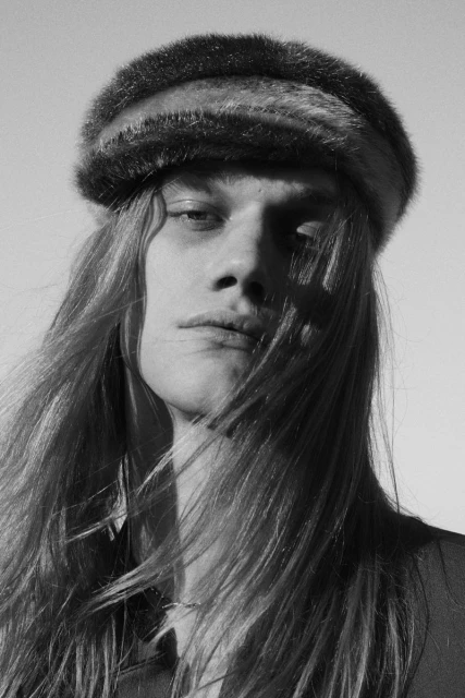 Black and white portrait of a young adult with long hair and a fur hat against a bright, cloudless sky.
