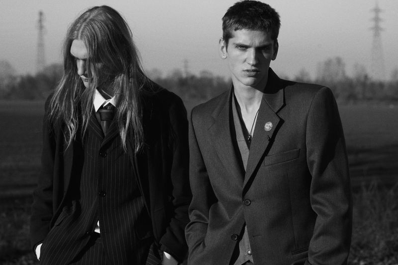 Two male models in dark tailored suits standing outdoors in an open landscape with power pylons visible in the hazy distance.