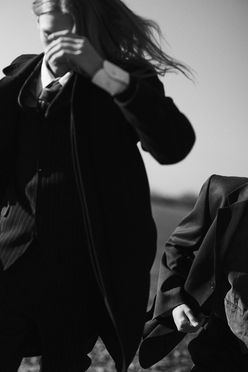Black and white photo of a long-haired model running in a dark overcoat and suit beside another tailored figure outdoors.