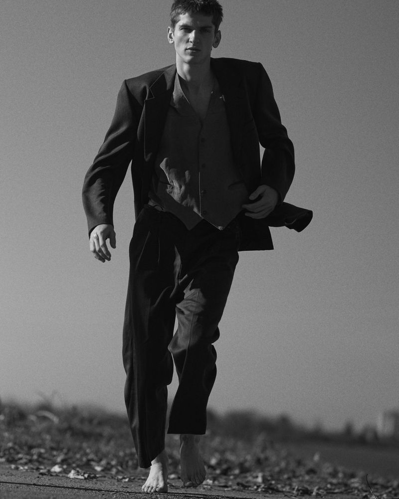 Man running barefoot wearing a tailored dark suit jacket and trousers against an outdoor sky background