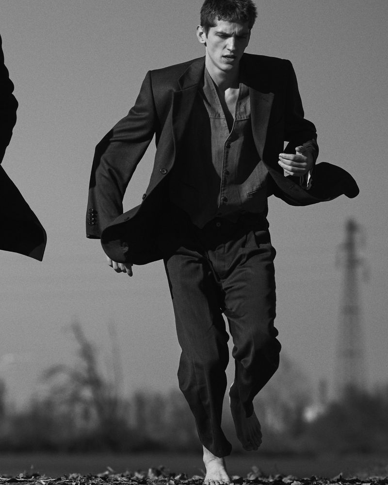 Black and white image of a barefoot man in a suit running in an open, gritty landscape with industrial structures in the background.