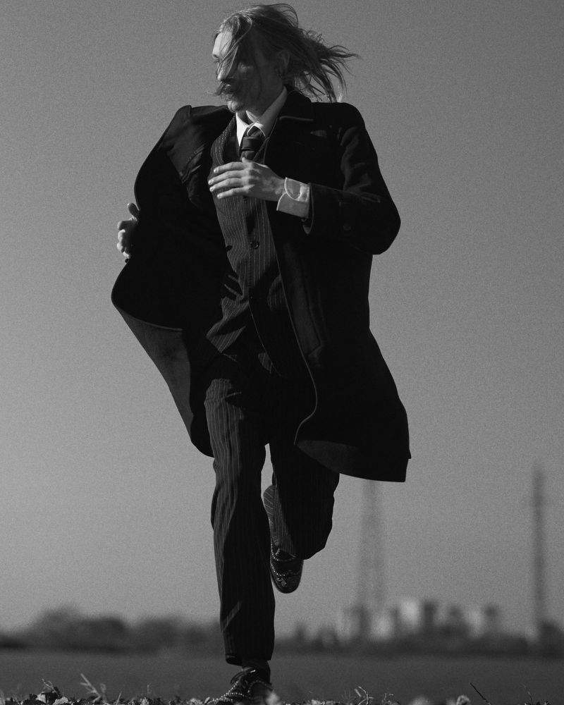 A man running forward through an open field wearing a dark tailored pinstripe suit and an open flowing overcoat.