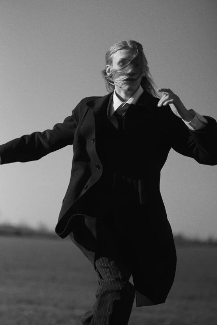 Black and white image of a long-haired person in a suit running in a field against a clear sky, hair covering face, open arms.