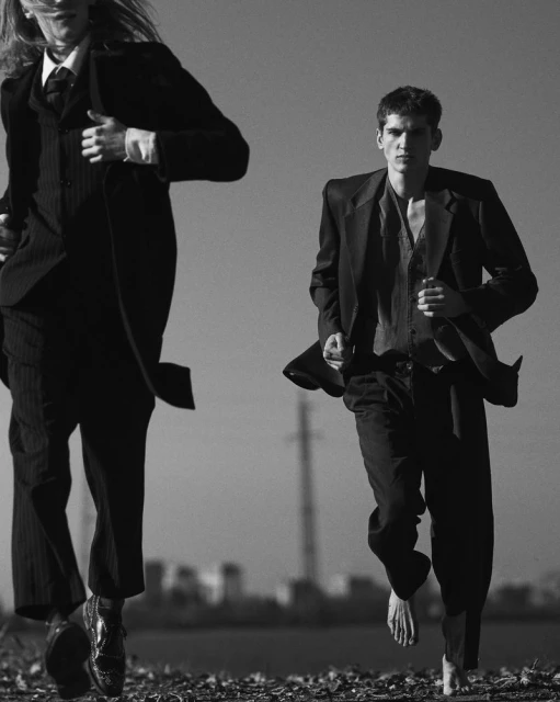 Two adult males running outdoors in suits against a clear sky and cityscape backdrop; one is barefoot, fashion editorial style in black and white.
