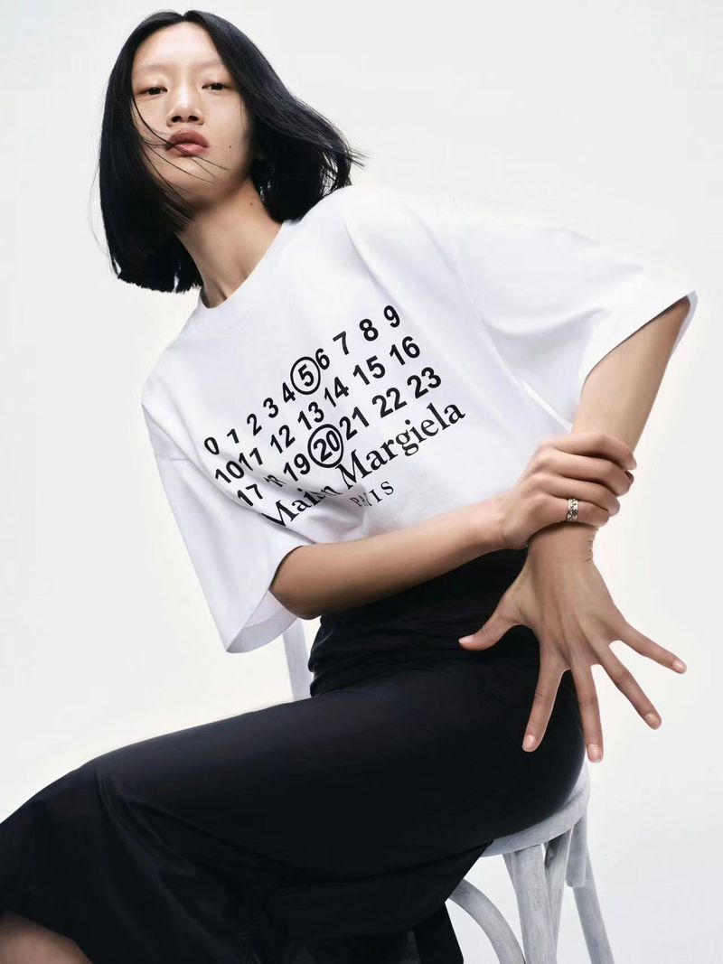 An adult woman is sitting while posing for the camera. She has short black hair and is wearing a white t-shirt and black skirt.