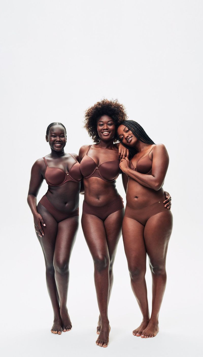 Three Black women in bras and underwear smiling in a studio setting with a white backdrop, showcasing body positivity and diverse beauty.