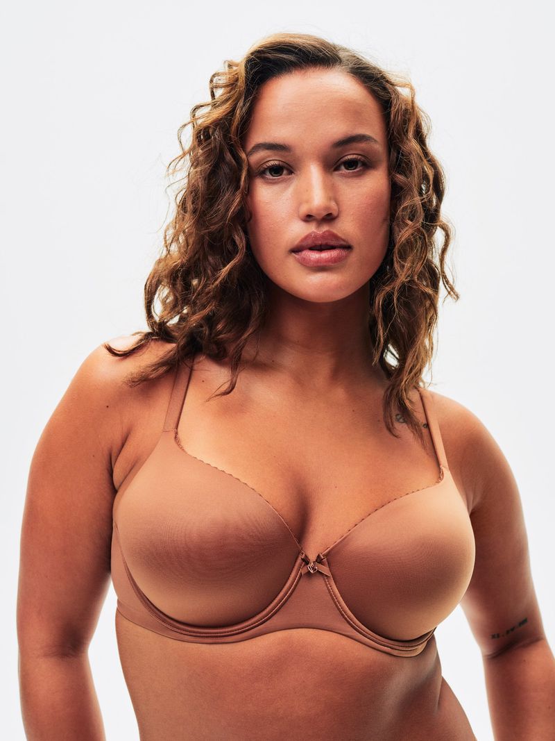 A woman with curly brown hair poses in front of a white background wearing a tan bra looking directly at the camera.