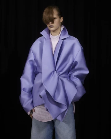 A person with long brown bangs poses in an oversized lavender coat and blue jeans against a dark backdrop in a studio-lit fashion shot.