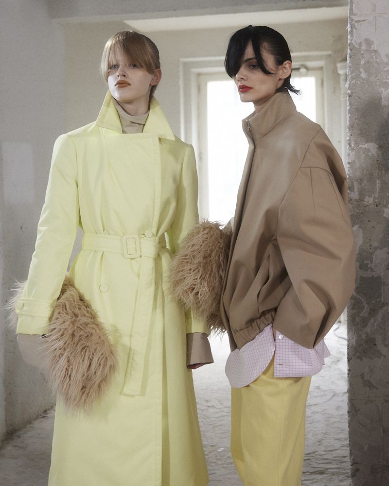 Two fair-skinned adult women in a sparse interior, wearing fashionable outerwear: a yellow coat with fur sleeves and a tan jacket.