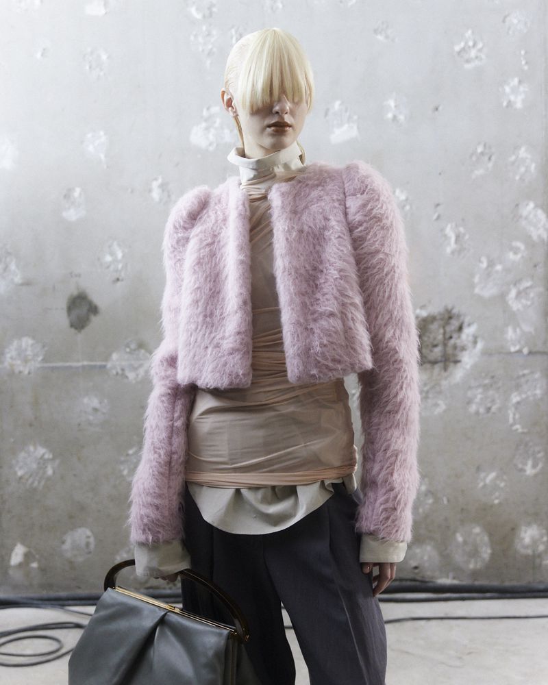 An adult woman with bleached hair and bangs wears a pink faux fur jacket with gray pants, holding a dark handbag against a textured wall.