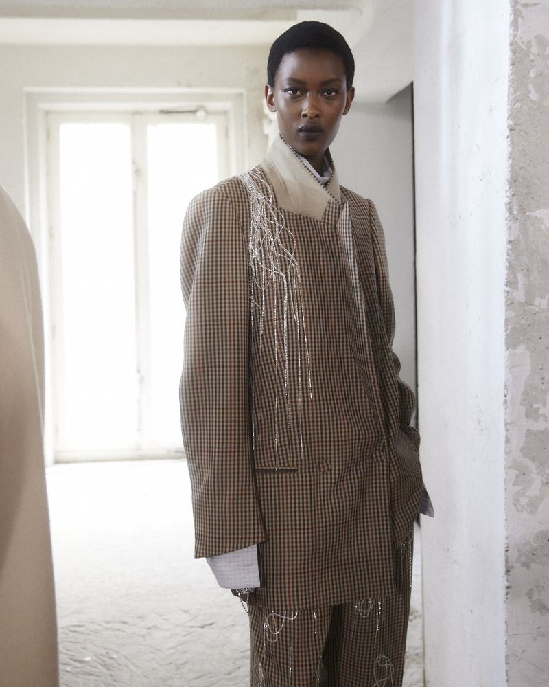 Fashionable woman in a tailored brown plaid suit posing indoors with neutral-tone walls. Statement garment for editorial or runway.