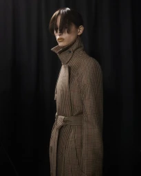 Model wearing a structured brown checkered coat with a high collar and belt against a dark backdrop