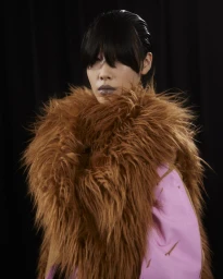 Model wearing a brown fluffy faux fur collar and pink garment against a dark background for Dries Van Noten