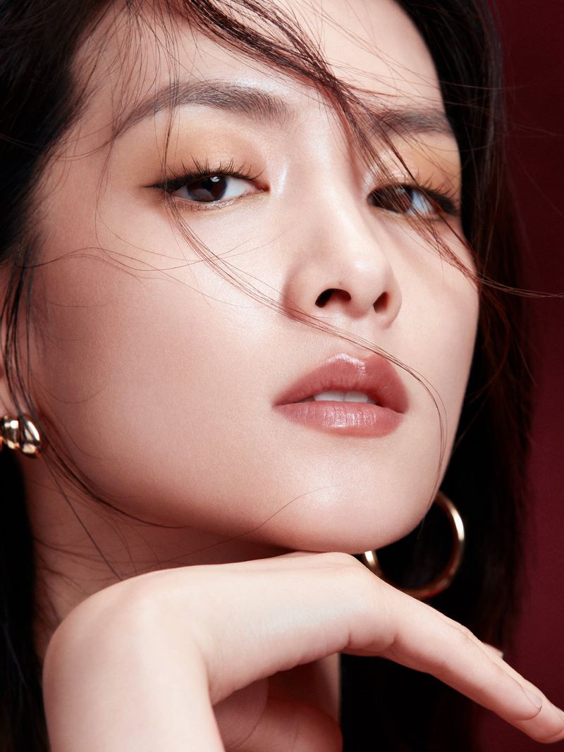 Close-up beauty shot of an adult woman with dark hair, gold earrings, brown eye shadow, and glossy lips against a dark red background.