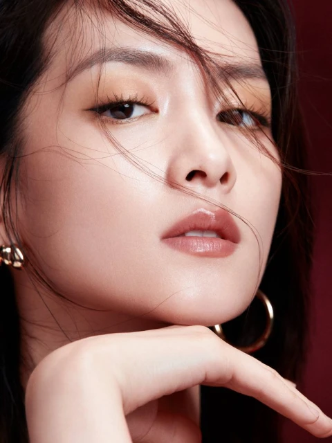 Close-up beauty shot of an adult woman with dark hair, gold earrings, brown eye shadow, and glossy lips against a dark red background.