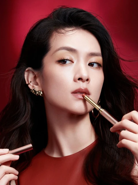 Close-up of an Asian woman applying coral lipstick with a gold applicator, set against a rich red background in a beauty setting.
