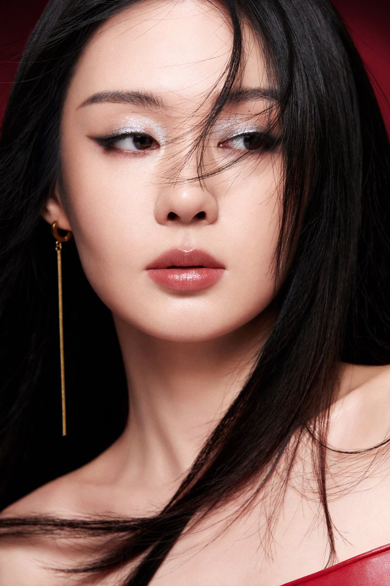 Close-up studio portrait of a young Asian woman with long black hair, shimmering eye makeup, gold dangle earring, against a red background.