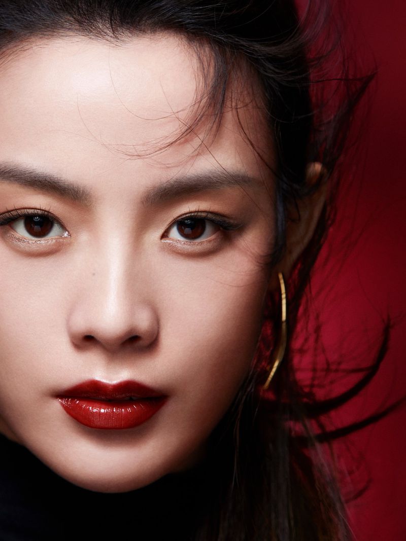 Close-up portrait of a woman with dark hair, brown eyes, red lipstick, and gold earrings against a dark red background.