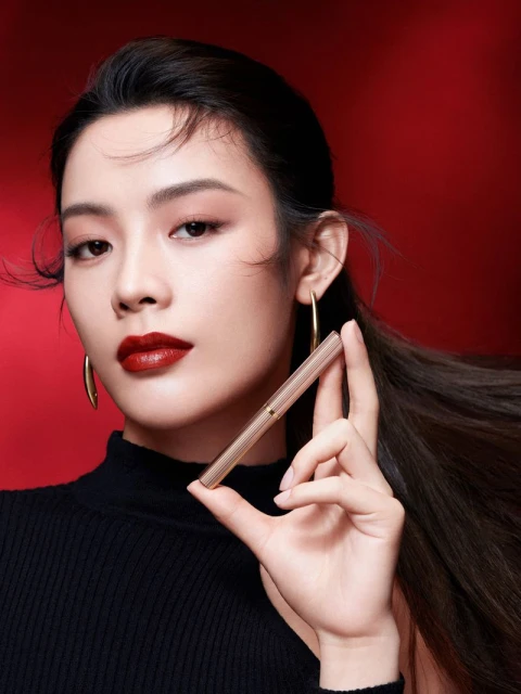Close-up of a woman with dark red lips, wearing gold hoop earrings and holding a gold lipstick tube against a red background.