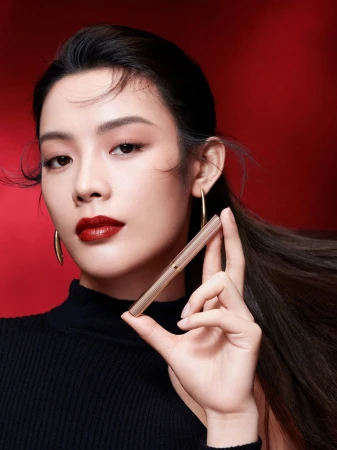 Close-up of a woman with dark red lips, wearing gold hoop earrings and holding a gold lipstick tube against a red background.