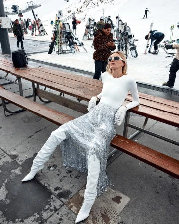 A woman in a white top and sequined skirt sits on a bench, wearing white boots, with snowy mountains and skiers in the background.