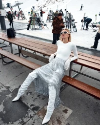 Model wearing a white top, shimmering skirt, and white boots seated on a wooden picnic table at a snowy ski resort.