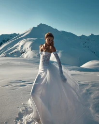 A woman in a white strapless gown standing in a vast snowy mountain landscape under a clear sky.