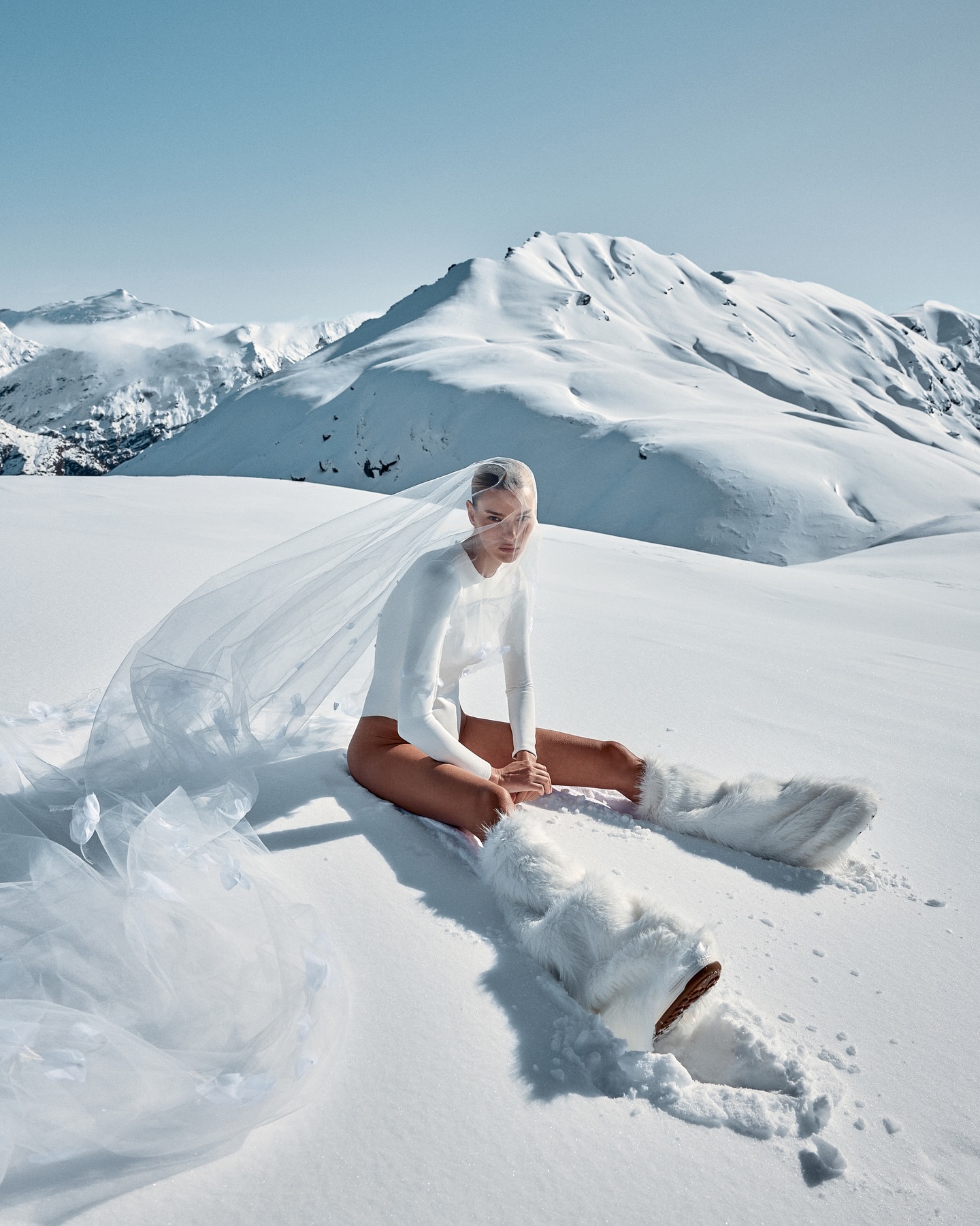 Woman in a white outfit and furry boots sits in a snowy mountain landscape with a long veil trailing behind her, under a clear blue sky.