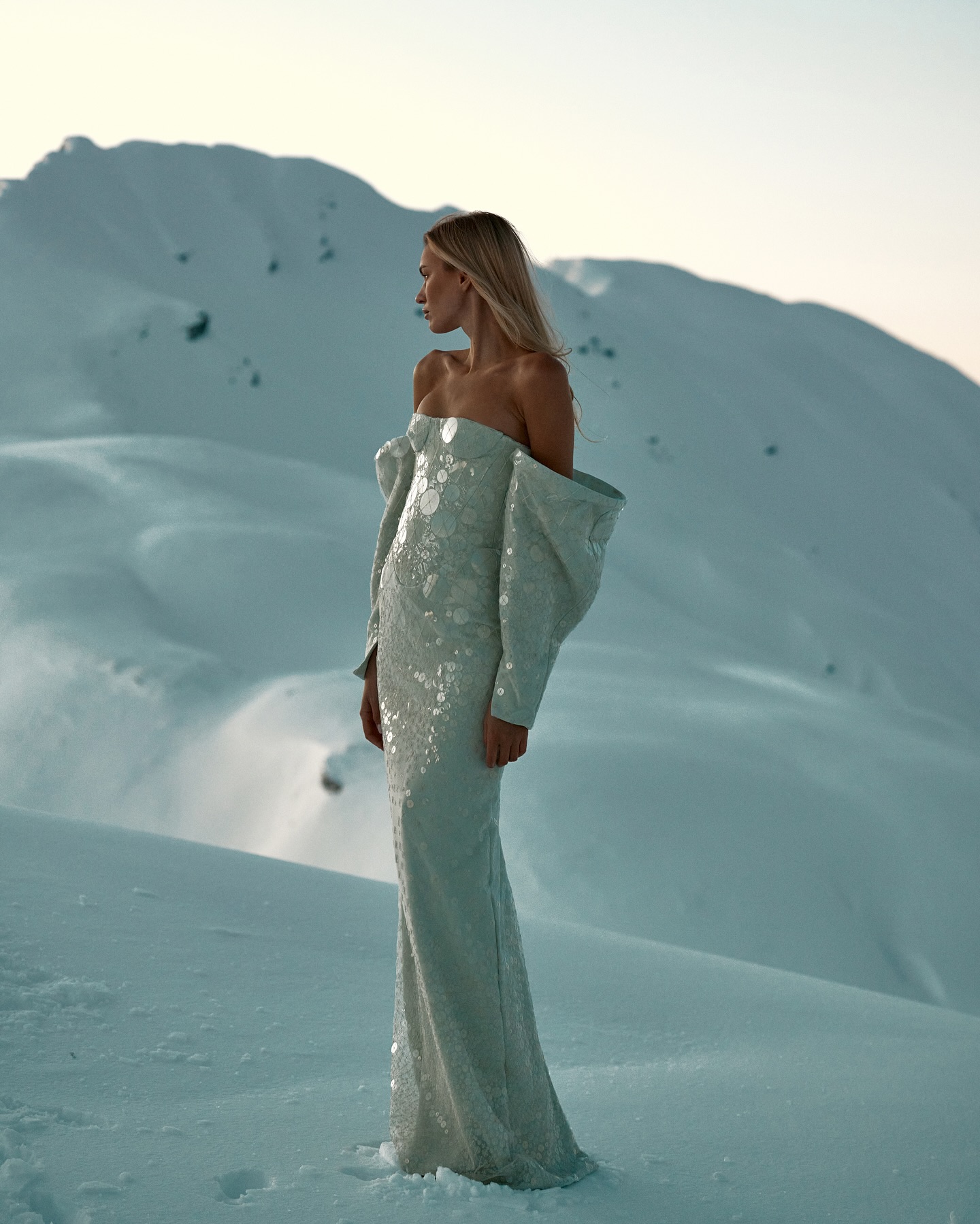 Model standing on a snowfield wearing a strapless pale dress against snowy mountains at sunset.