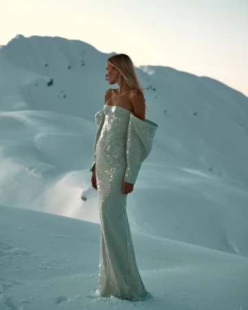 Model standing on a snowfield wearing a strapless pale dress against snowy mountains at sunset.