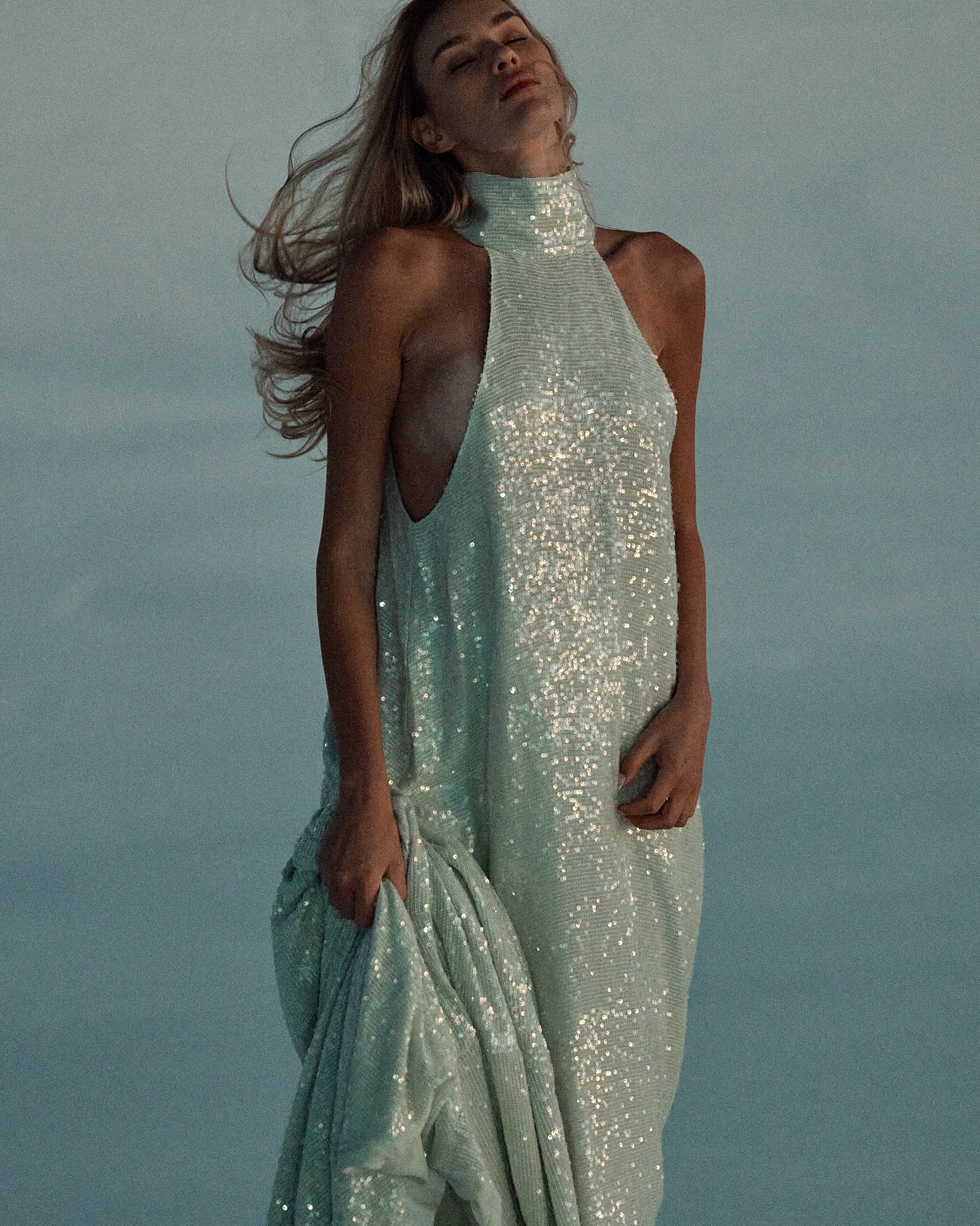 Model wearing a shimmering high-neck sequin gown standing against an open sky background