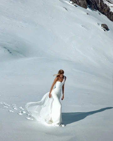 Model in a long white bridal gown walking across a snowy mountain slope