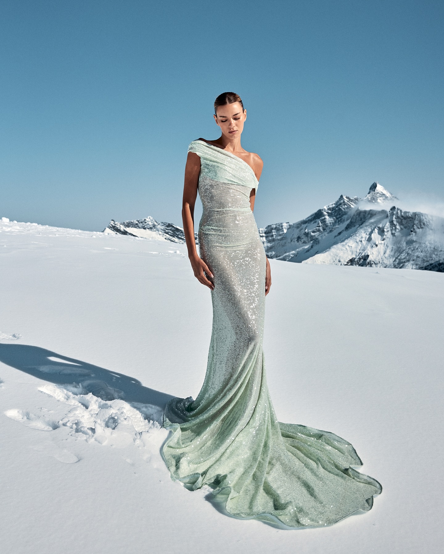A fashion model poses in a shimmering gown in a snow landscape with mountains in the background, under a clear blue sky.