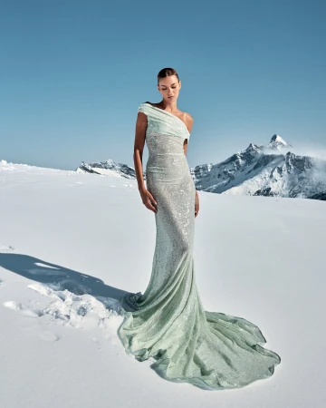 A fashion model poses in a shimmering gown in a snow landscape with mountains in the background, under a clear blue sky.