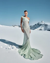 Model wearing a glittering pale green evening gown standing on a snowfield against snowy mountain peaks under a clear blue sky