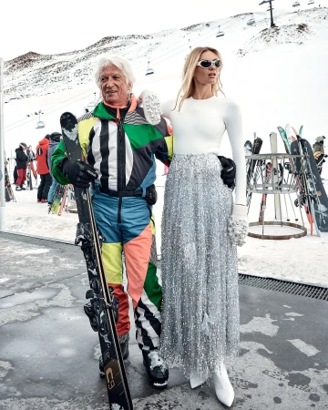 Fashion photo of a man with skis and a woman in a sequined skirt posing on a snowy slope with ski lifts in the background.