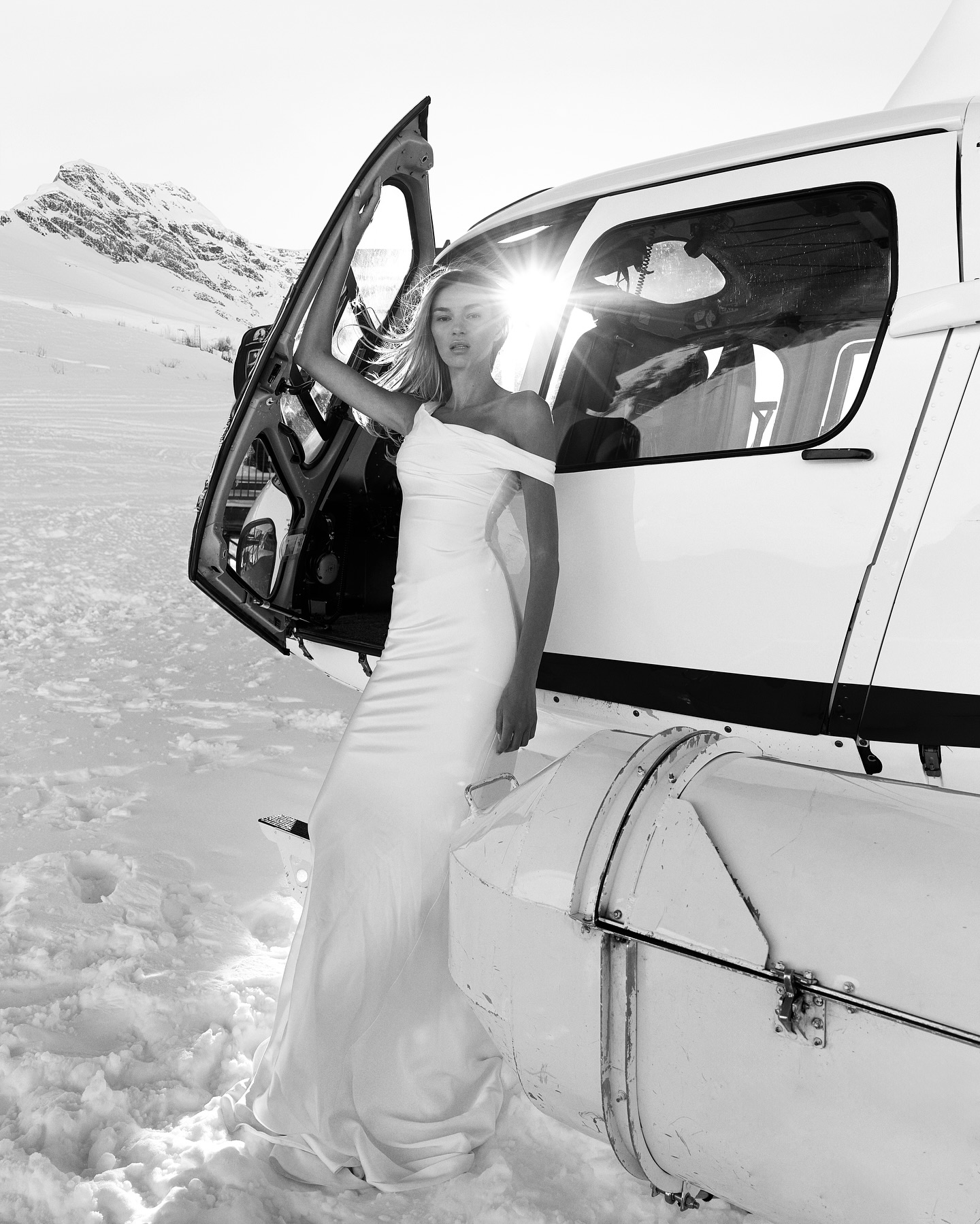 A woman in a white dress exits a helicopter on a snowy mountain with sunlight and shadows, showcasing fashion in an outdoor setting.
