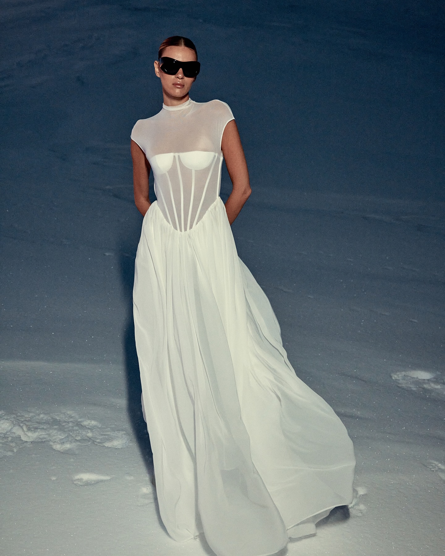 A woman in a long white dress and sunglasses stands on snow, creating a striking contrast between fashion and the natural environment.