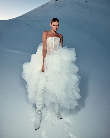 Woman in a white dress and boots poses in a snowy landscape. A mountain rises in the background under a clear sky, creating a striking contrast.