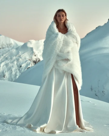 A woman stands on a snow-covered mountain wearing a white dress and a white faux fur shawl, creating a striking winter fashion statement.