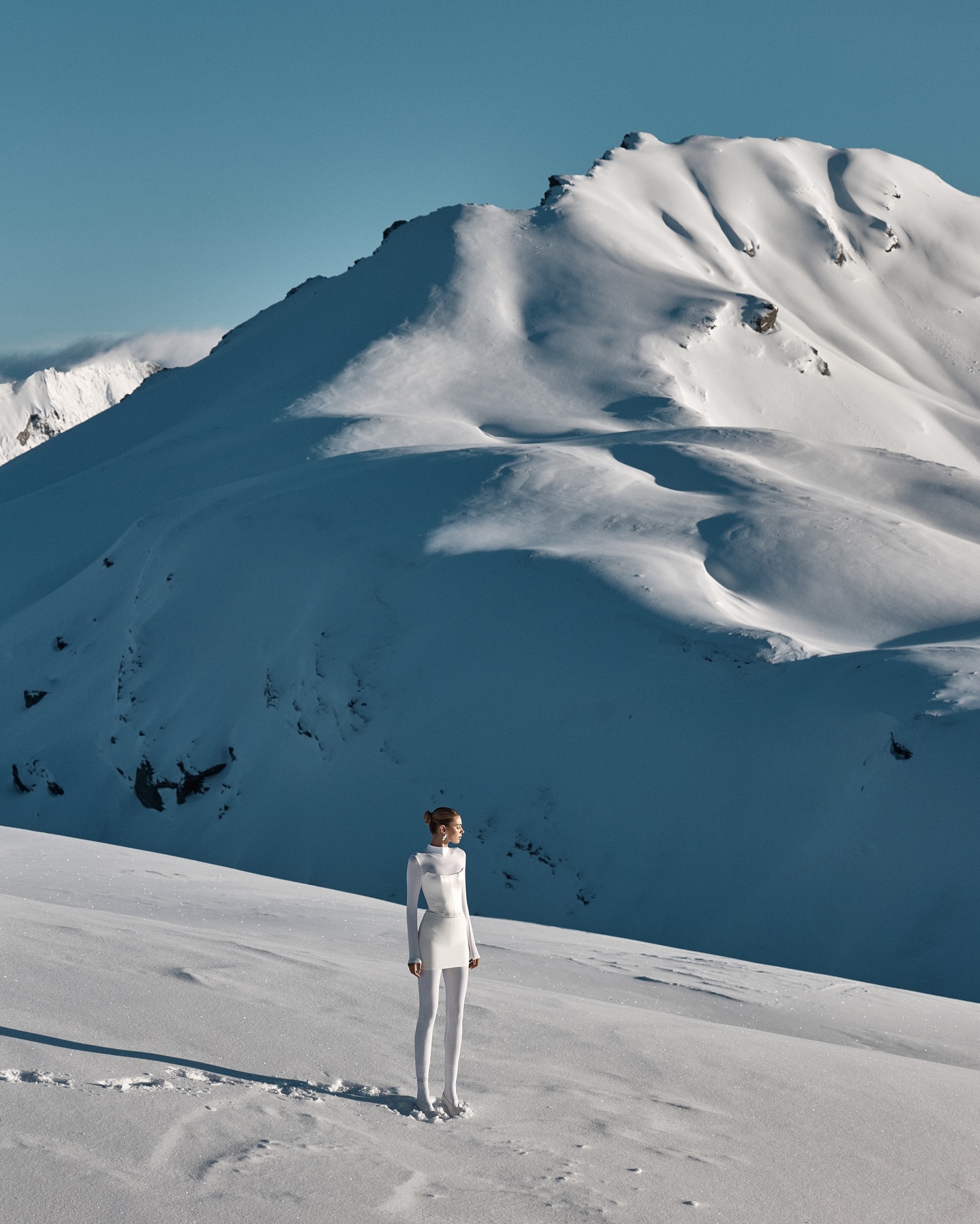 A woman in a white outfit stands on a snow-covered hill, with a large, snow-covered mountain range stretching behind her under a clear blue sky.