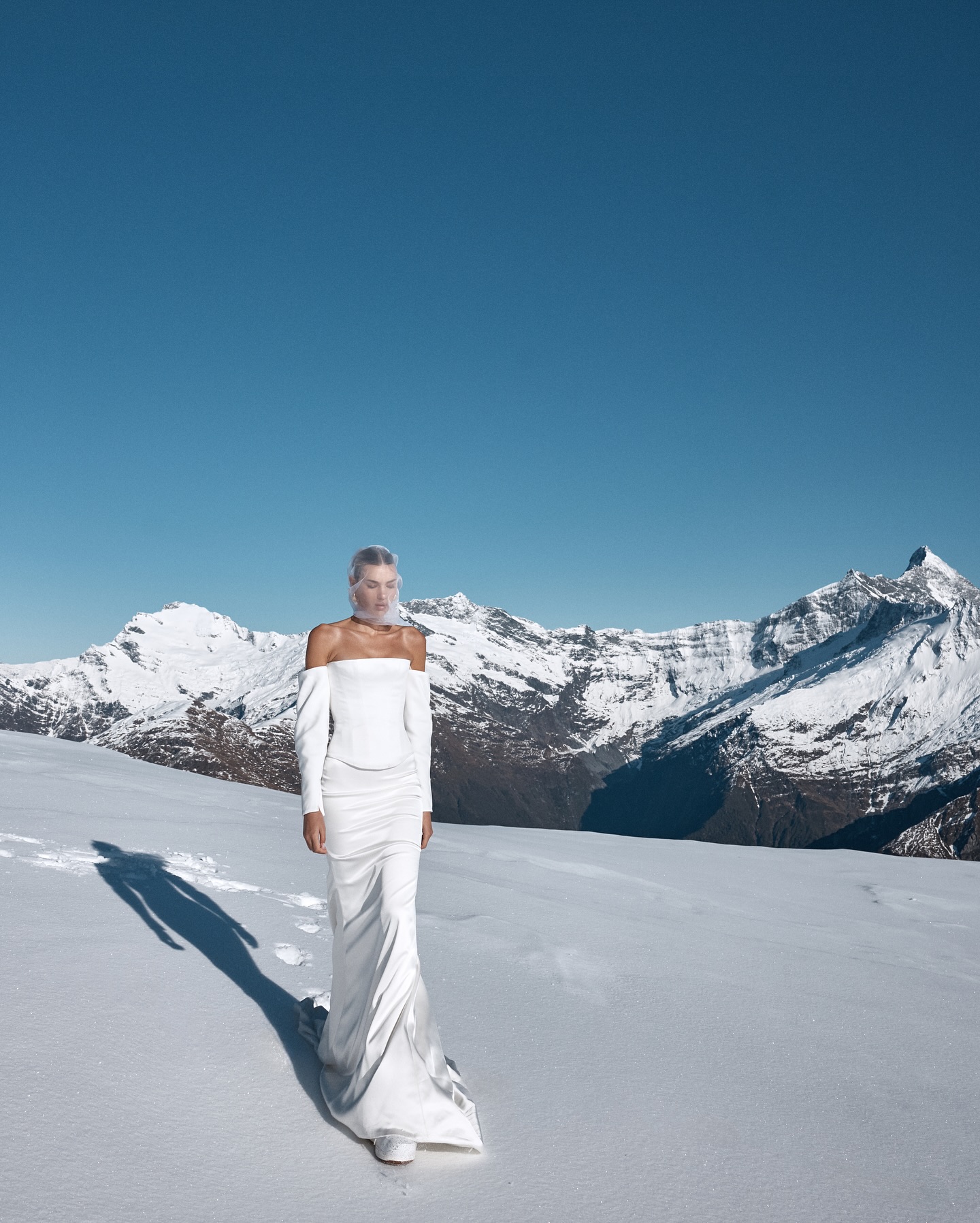 A woman in a white dress walks on a snow-covered landscape, with a clear blue sky and snow-capped mountains in the background on a sunny day.